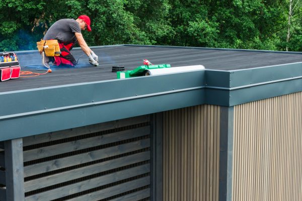 A worker is applying protective coating to a flat roof of a contemporary building surrounded by lush greenery during a bright day.